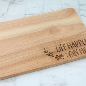 May include: A wooden cutting board with the text "Life Happens...Gin Helps" engraved on it. The text is surrounded by a small floral design.