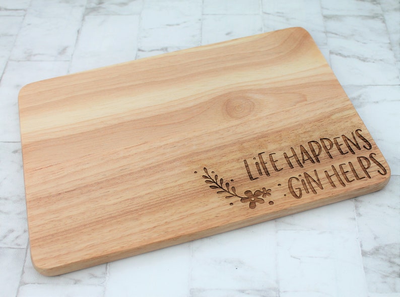 May include: A wooden cutting board with the text "Life Happens...Gin Helps" engraved on it. The text is surrounded by a small floral design.
