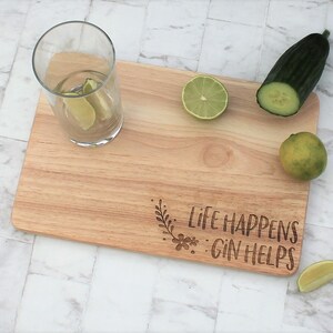May include: Wooden cutting board with the text "Life Happens: Gin Helps" engraved on it. There are limes, a cucumber, and a glass of water with lime wedges on the board.