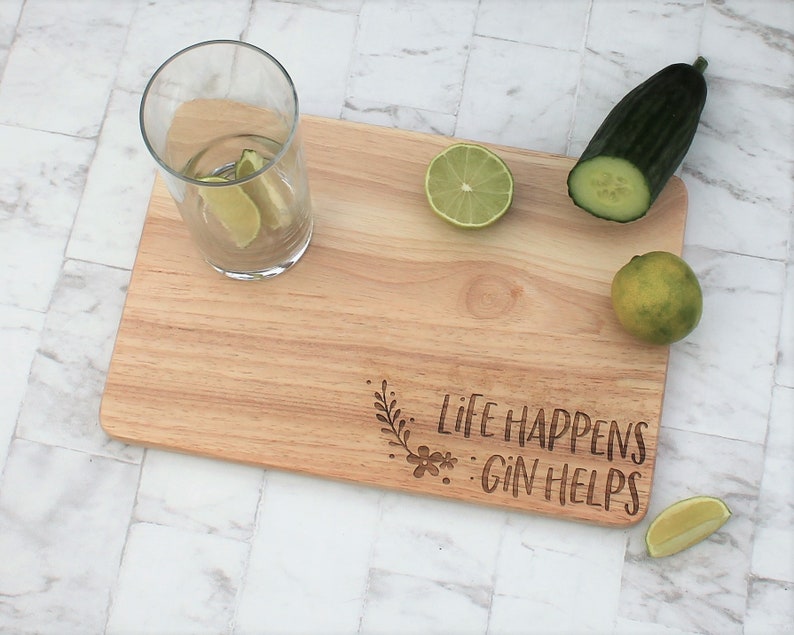 May include: Wooden cutting board with the text "Life Happens: Gin Helps" engraved on it. There are limes, a cucumber, and a glass of water with lime wedges on the board.