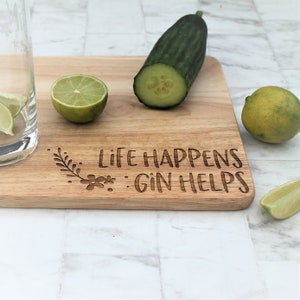 May include: A wooden cutting board with the text "Life Happens Gin Helps" engraved on it. There are limes, a cucumber, and a glass of gin on the board.