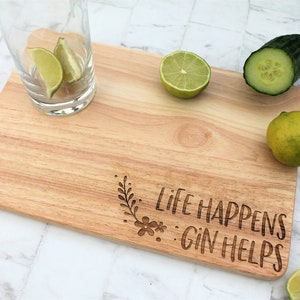 May include: A wooden cutting board with the text "Life Happens Gin Helps" engraved on it. The board is surrounded by limes and cucumbers.