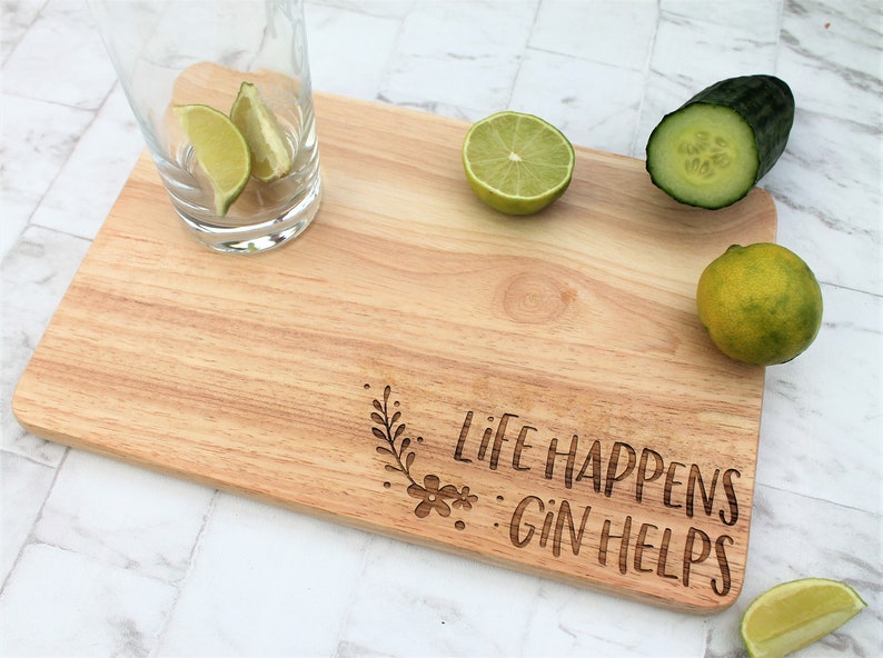 May include: A wooden cutting board with the text "Life Happens Gin Helps" engraved on it. The board is surrounded by limes and cucumbers.