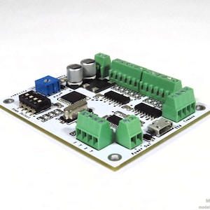 May include: A white circuit board with various electronic components, including green terminal blocks, black integrated circuits, and a blue potentiometer. The board has text that reads "USB Connex" and "Power Out".