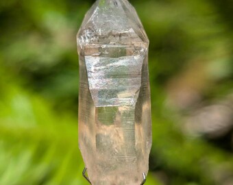 May include: A clear, elongated quartz crystal point with a pointed termination and natural horizontal striations along its vertical facets. The crystal is held upright by a silver-toned metal wire stand that grips the base of the mineral specimen.