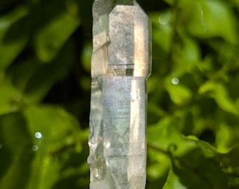 May include: A clear, elongated quartz crystal point with a hexagonal structure and natural facets. The crystal is held upright by a silver-toned metal pronged base, showcasing internal clarity and light reflections throughout its transparent body.