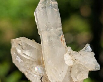 May include: A cluster of clear, translucent quartz crystals with a prominent central point and smaller surrounding formations. The mineral specimen is held by a metallic, textured mechanical pencil tip, showcasing natural facets and internal inclusions.