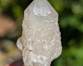 May include: A clear, translucent quartz crystal point with a textured, jagged surface and a pointed termination. The mineral specimen features natural crystalline formations and small, irregular facets across its body, displaying a white and icy appearance.
