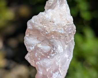 May include: A raw, translucent pale pink and white mineral specimen with a botryoidal, bumpy surface texture. The stone features irregular, rounded formations and a glossy, crystalline appearance throughout its jagged, organic, and uneven shape.