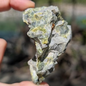 May include: A rough, irregularly shaped rock with a mix of gray and green hues. The rock has a crystalline structure with visible layers and textures. The image is taken outdoors, with a blurred background of trees and sky.