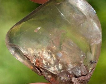 May include: A polished, translucent quartz crystal with a smooth, rounded shape. The stone features a clear, pale yellow body with visible internal fractures and a dark, textured mineral inclusion at the base, showing earthy brown and metallic tones.