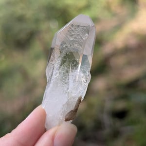 May include: A clear quartz crystal with a pointed tip and a rough, natural texture. The crystal is held up against a blurred green background, showcasing its translucent quality and natural form. A small, brown inclusion is visible.