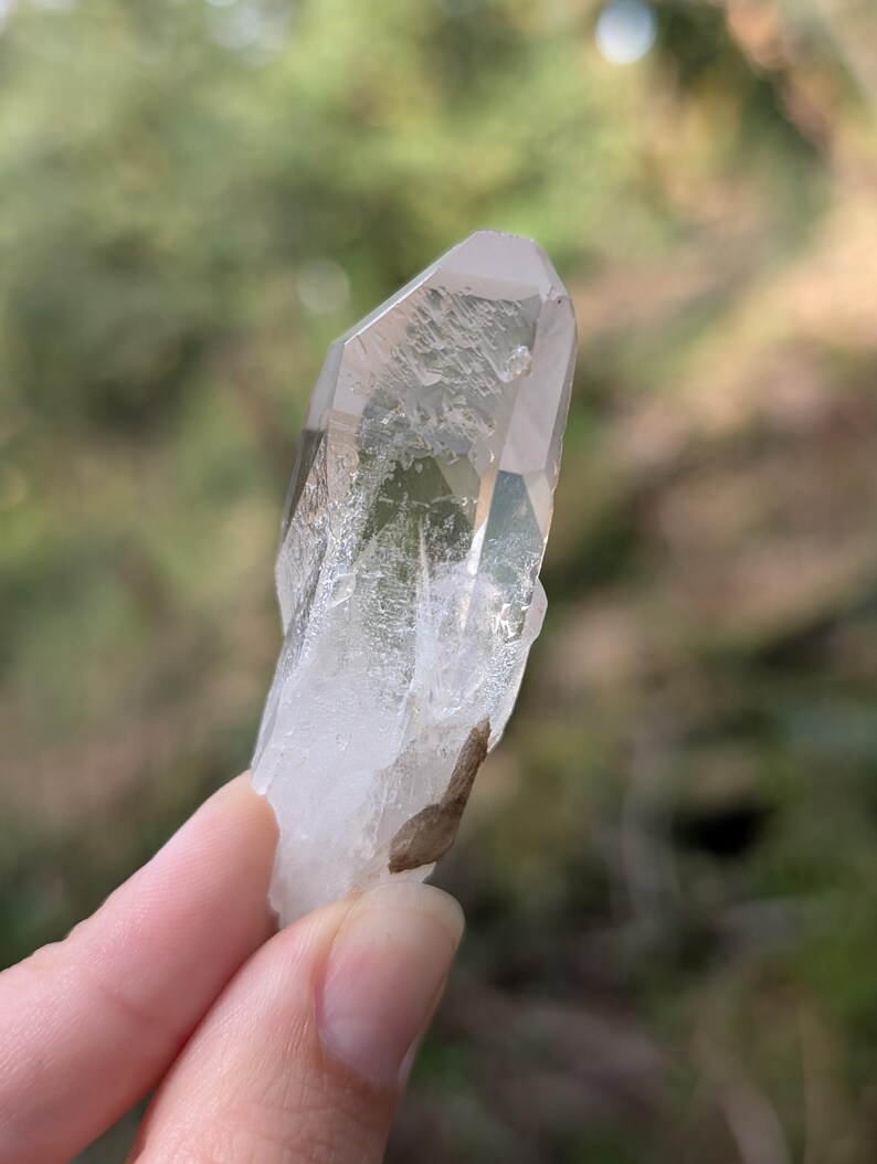 May include: A clear quartz crystal with a pointed tip and a rough, natural texture. The crystal is held up against a blurred green background, showcasing its translucent quality and natural form. A small, brown inclusion is visible.