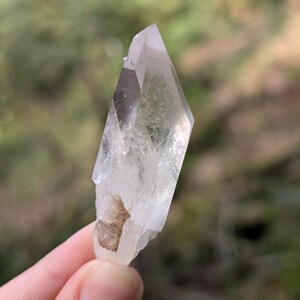 May include: A clear quartz crystal with a pointed tip and a rough, natural base. The crystal is held against a blurred green background, showcasing its translucent quality and natural form. The crystal's facets catch the light, highlighting its clarity.
