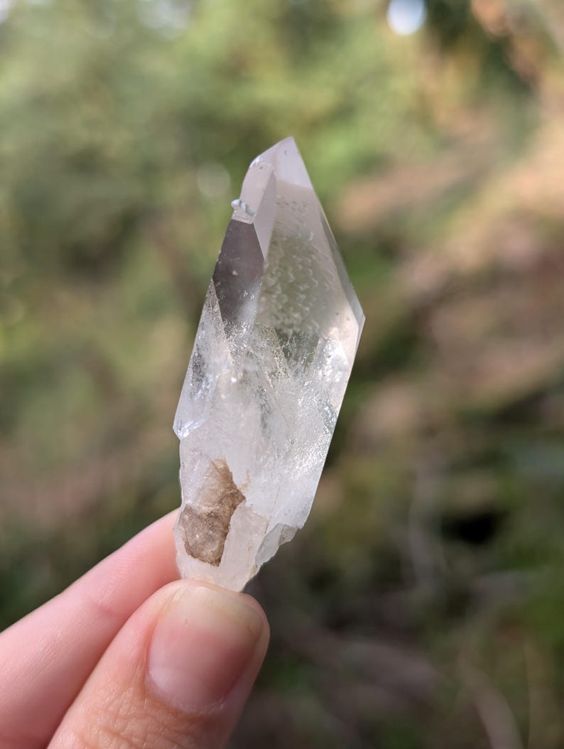 May include: A clear quartz crystal with a pointed tip and a rough, natural base. The crystal is held against a blurred green background, showcasing its translucent quality and natural form. The crystal's facets catch the light, highlighting its clarity.