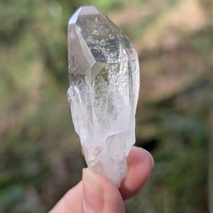 May include: A clear quartz crystal with a faceted top and a rough, textured base. The crystal is held against a blurred green background, showcasing its natural clarity and shape. The crystal is a light, translucent color.