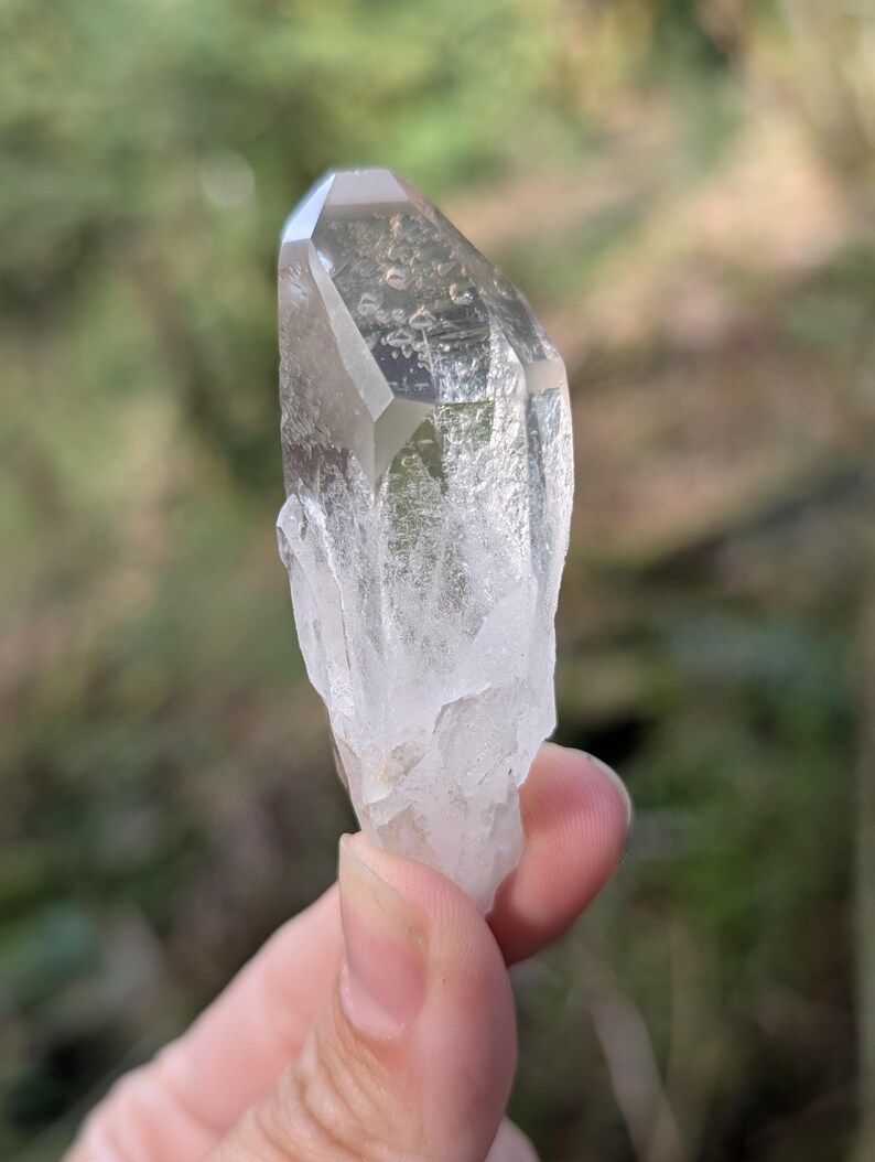 May include: A clear quartz crystal with a faceted top and a rough, textured base. The crystal is held against a blurred green background, showcasing its natural clarity and shape. The crystal is a light, translucent color.