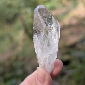 May include: A clear quartz crystal with a faceted top and a rough, textured base. The crystal is held against a blurred green background, showcasing its natural clarity and form. The quartz is a light, translucent color.