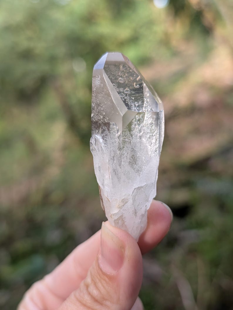 May include: A clear quartz crystal with a faceted top and a rough, textured base. The crystal is held against a blurred green background, showcasing its natural clarity and form. The quartz is a light, translucent color.