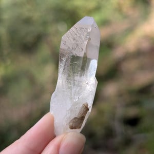 May include: A clear quartz crystal with a pointed tip and a rough, textured surface. The crystal has a natural, organic shape and a small, brown inclusion near the base. The background is blurred, with green and brown tones.