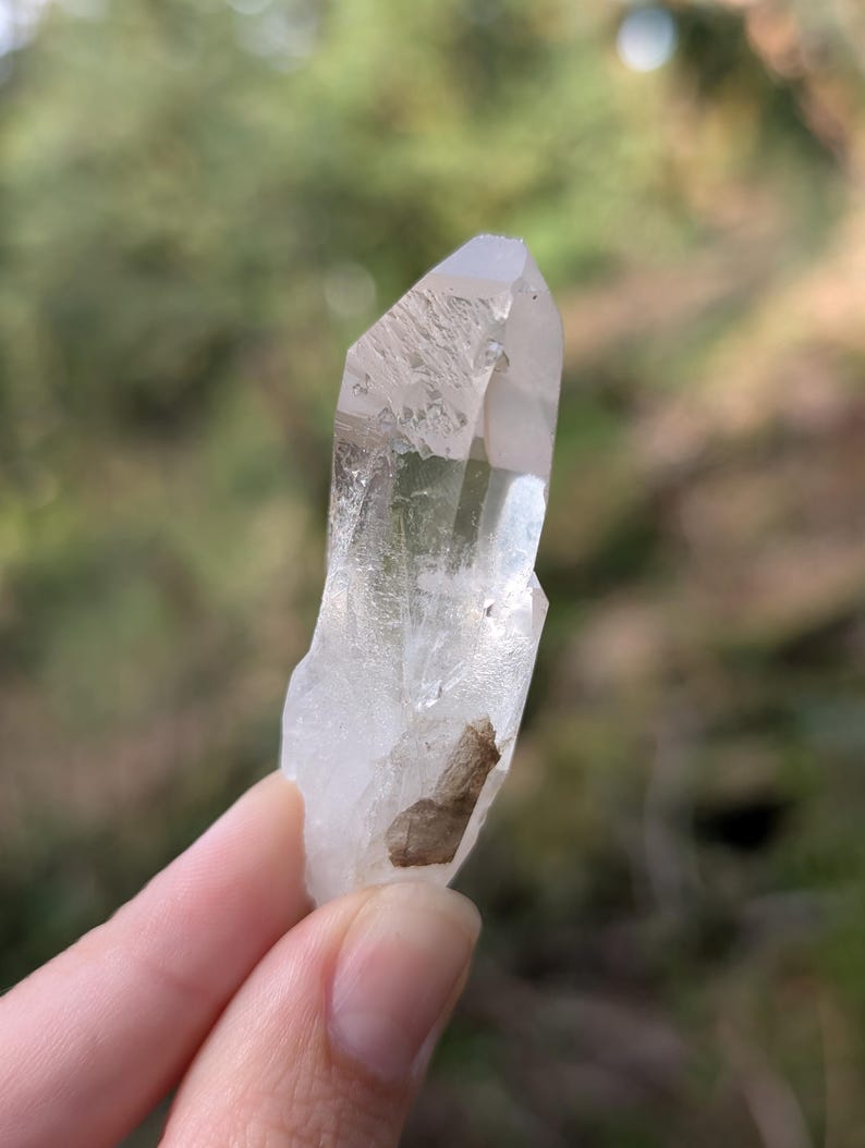 May include: A clear quartz crystal with a pointed tip and a rough, textured surface. The crystal has a natural, organic shape and a small, brown inclusion near the base. The background is blurred, with green and brown tones.