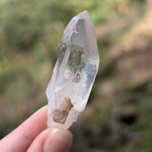 May include: A clear quartz crystal with a pointed tip and a rough, natural base. The crystal is held up against a blurred green background, showcasing its translucent quality and natural inclusions. The crystal is approximately 3 inches long.