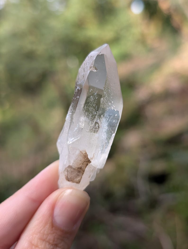 May include: A clear quartz crystal with a pointed tip and a rough, natural base. The crystal is held up against a blurred green background, showcasing its translucent quality and natural inclusions. The crystal is approximately 3 inches long.