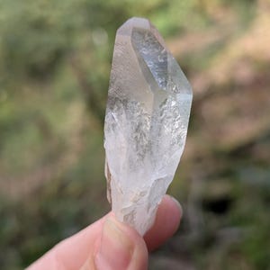 May include: A clear quartz crystal with a pointed tip and a rough, textured base. The crystal is held against a blurred green and brown background, showcasing its natural clarity and form. The crystal is approximately 3 inches in length.