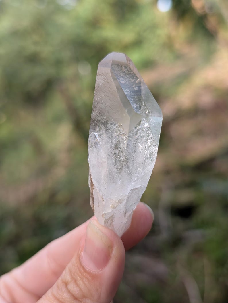 May include: A clear quartz crystal with a pointed tip and a rough, textured base. The crystal is held against a blurred green and brown background, showcasing its natural clarity and form. The crystal is approximately 3 inches in length.