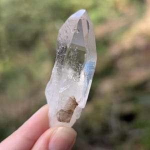 May include: A clear quartz crystal with a pointed tip. The crystal is held against a blurred green background. The quartz has a natural, rough texture and a small brown inclusion near the base. The crystal is approximately 3 inches long.