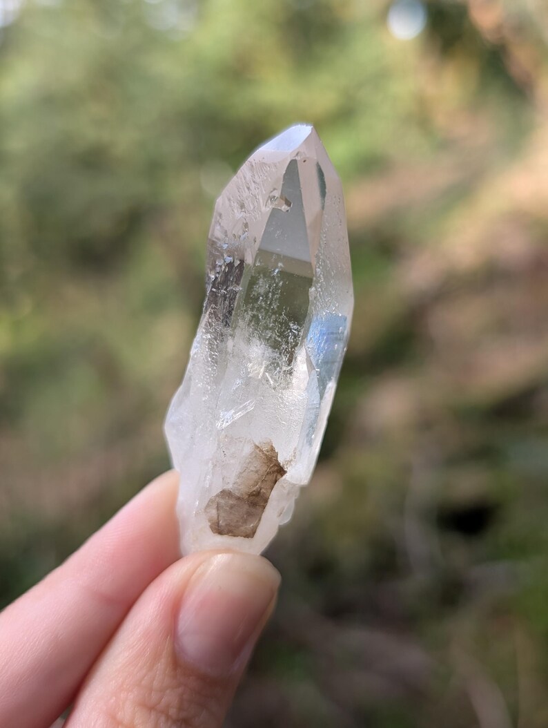 May include: A clear quartz crystal with a pointed tip. The crystal is held against a blurred green background. The quartz has a natural, rough texture and a small brown inclusion near the base. The crystal is approximately 3 inches long.