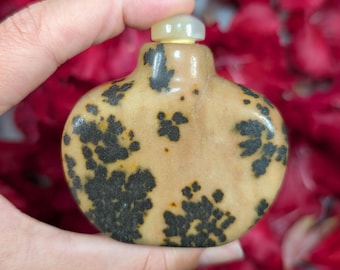 May include: A small, antique-style bottle with a rounded shape and a light brown base. The bottle is adorned with dark, irregular spots. It has a small, light-colored stopper. The background is a blur of red.