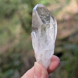 May include: A clear quartz crystal with a faceted top and a rough, textured base. The crystal is held against a blurred green background, showcasing its natural clarity and form. The crystal is a light, translucent color.