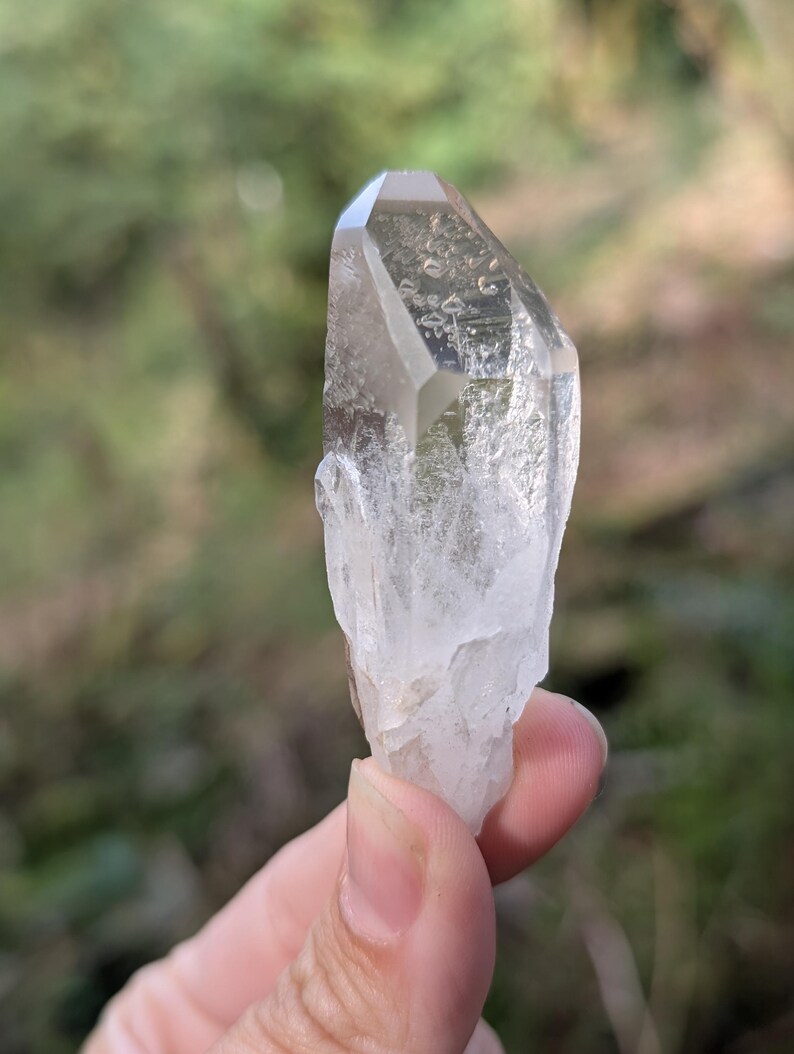 May include: A clear quartz crystal with a faceted top and a rough, textured base. The crystal is held against a blurred green background, showcasing its natural clarity and form. The crystal is a light, translucent color.