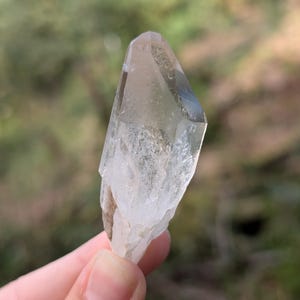 May include: A clear quartz crystal with a pointed tip and a rough, textured base. The crystal is held against a blurred green background, showcasing its natural, translucent appearance. The crystal is approximately 3 inches in length.