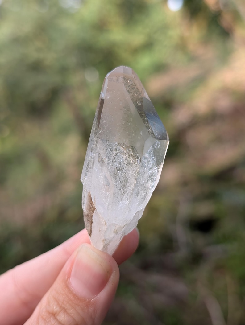 May include: A clear quartz crystal with a pointed tip and a rough, textured base. The crystal is held against a blurred green background, showcasing its natural, translucent appearance. The crystal is approximately 3 inches in length.