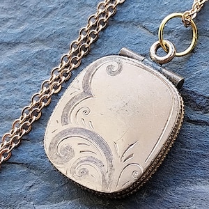 May include: A gold-toned, rectangular locket pendant with an ornate, engraved design on the front. The locket hangs from a delicate gold chain. The background is a textured, dark blue-gray surface.