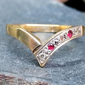 May include: A gold-toned V-shaped ring with a decorative band. The band features small, round, red gemstones and clear, square-cut stones. The ring is set against a blue-gray surface.