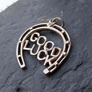 May include: A silver-colored horseshoe pendant with the words "GOOD LUCK" in the center. The horseshoe has a loop at the top for a chain. The pendant is on a dark gray surface.