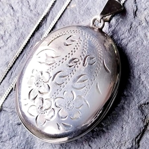 Könnte beinhalten: Ovaler silberner Medaillon-Anhänger mit floralen und Blattgravuren. Das Medaillon hängt an einer filigranen Silberkette. Der Anhänger hat ein Scharnierdesign und eine polierte Oberfläche. Das florale Design ist in die Oberfläche des Medaillons geätzt.