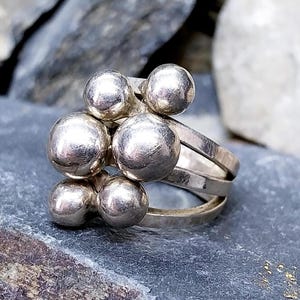 May include: A silver ring featuring a cluster of six round, polished spheres. The ring has a multi-band design, with three parallel bands forming the base. The spheres are arranged in a visually appealing, asymmetrical pattern.