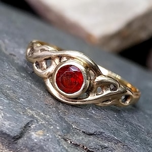 Puede incluir: Un anillo dorado con un diseño ornamentado. El anillo presenta una gema roja, redonda y facetada, engastada en el centro. La banda tiene un patrón calado. El anillo se muestra sobre una superficie gris texturizada.