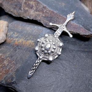May include: A silver-toned brooch shaped like a sword with a circular shield attached to the center. The shield features a central raised dome surrounded by smaller spherical accents. The sword hilt and blade have intricate engraved details.