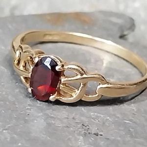 Op de afbeelding: Een gouden ring met een ovale, dieprode edelsteen. De ring heeft een delicaat, verweven ontwerp op de band, en de edelsteen is gezet in een vier-klauwzetting. De ring wordt gepresenteerd op een grijze ondergrond.