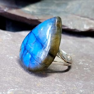 Puede incluir: Anillo de plata con una gran piedra de labradorita pulida. La piedra tiene forma de lágrima y muestra colores azul brillante e iridiscente con vetas oscuras. El anillo está sobre una superficie gris.
