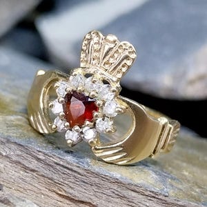 May include: A gold Claddagh ring featuring a central heart-shaped red gemstone surrounded by a halo of small clear stones. The ring design includes two hands holding the heart and a crown positioned above it, all crafted in a polished gold metal.