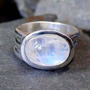 Puede incluir: Un anillo de plata con una gran piedra lunar ovalada. La piedra lunar tiene un aspecto lechoso e iridiscente, con destellos azules y blancos. La banda del anillo es gruesa y tiene un diseño simple y moderno. El anillo está sobre una superficie oscura y texturizada.