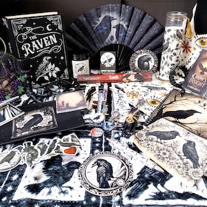 Könnte beinhalten: Eine Sammlung von Raben-Artikeln, darunter ein Buch The Raven, ein Fächer, ein Crows Insight Kartendeck, eine Kerze, ein Spiegel, Aufkleber und Schreibwaren. Die Gegenstände zeigen schwarz-weiße Illustrationen von Raben auf gemustertem Stoff.