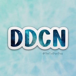 May include: A white sticker with the letters "DDCN" in a blue and teal watercolor design. The sticker has a white border and is set against a light blue and teal tie-dye background. The text "@TheTieDyeDog" is at the bottom.