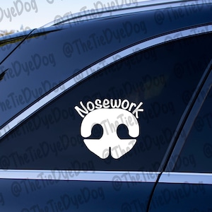 以下が含まれることがあります： 黒い車の窓に、犬の鼻のアイコンと「Nosework」という文字が書かれた白いビニールステッカー